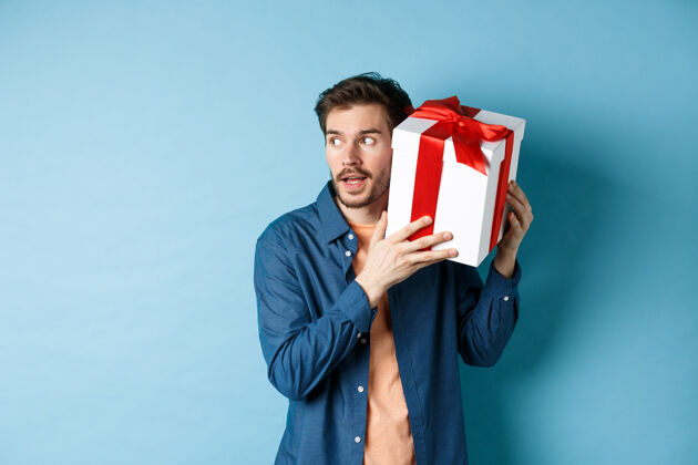 丈夫情人节好奇的男朋友拿着礼品盒靠近耳朵 试图猜出惊喜礼物里面是什么 站在蓝色的背景上微笑浪漫男人