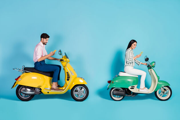 轻便摩托车全尺寸侧面照片正面两人妻子丈夫司机使用手机浏览互联网目的地旅行旅行驾驶摩托车隔离蓝色墙壁一起驾驶女孩