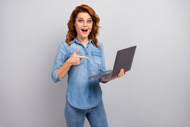 互联网惊讶女人的肖像使用笔记本电脑搜索无线连接显示广告促销印象深刻尖叫哇omg穿牛仔衬衫隔离灰色墙壁促销社交休闲