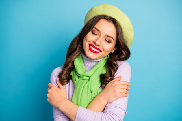 春天特写照片迷人的女士闭上眼睛拥抱自己和平内心和谐的概念穿绿色贝雷帽紫色套头衫围巾孤立的蓝色墙壁自我贝雷帽年轻