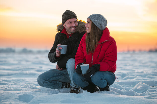 雪积极的年轻人穿着暖和的外套 拿着热水瓶坐在雪地上 在乡下喝着热饮料杯子活动田野