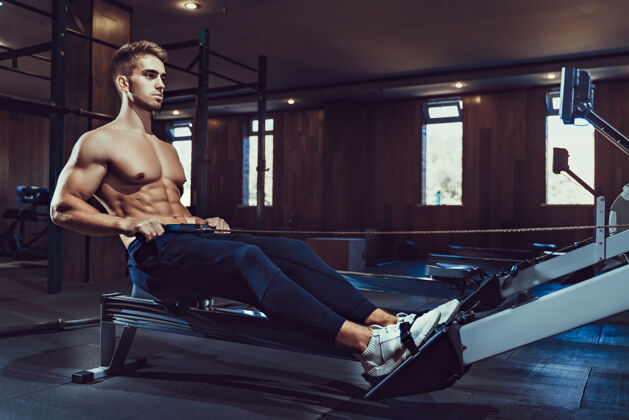 重量肌肉发达的健美运动员在运动服训练后坐在锻炼机上在黑暗的氛围中 在健身房里拥有完美健身身体的男人的侧视图健美 健身的概念健身房运动训练