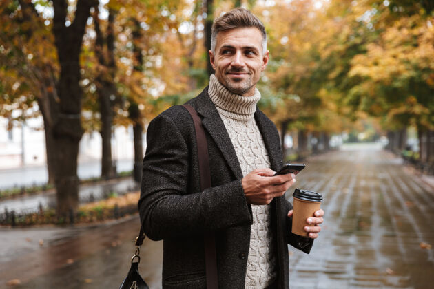 电话30多岁的帅哥穿着暖和的衣服 在户外穿过秋日公园 用手机拍照散步成人休闲