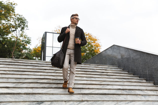 市区30多岁的欧洲男人戴着眼镜 穿过城市街道 手里拿着外卖咖啡中心胡须站立