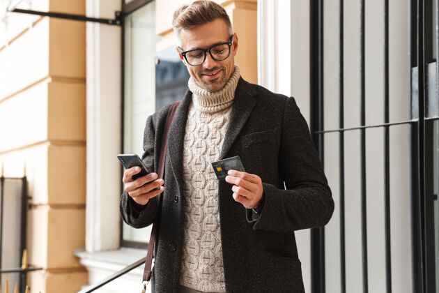 休闲帅哥穿着外套走在户外 用手机和信用卡在网上购物账单应用程序景点