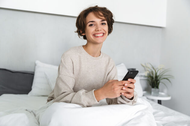 应用程序微笑的年轻女孩 早上在床上放松 用手机休闲连接黑发