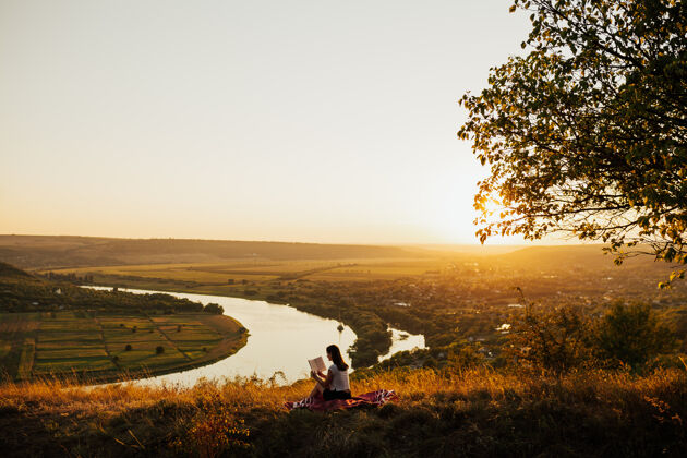 生活方式年轻的女游客坐在山上 以河流为背景 日落时在山上看书景点举行冒险