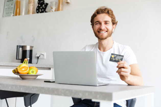学习快乐的小胡子男人在家里的桌子旁用手提电脑拿着信用卡聊天成人帅气休闲