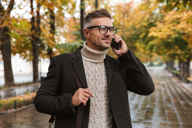 通话图为30多岁的快乐男子汉穿着暖和的衣服 在户外穿过秋天的公园 并使用手机公园天气时尚