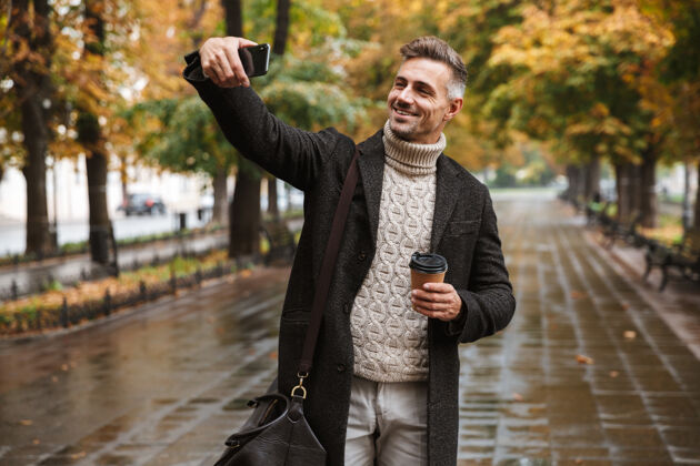 吸引力30多岁的快乐男人穿着暖和的衣服在户外穿过秋天公园 用手机自拍的照片林荫大道时尚享受