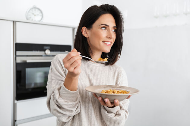 室内一位30多岁的成年妇女站在家里的现代厨房里吃着早餐中的玉米片水果满意消遣