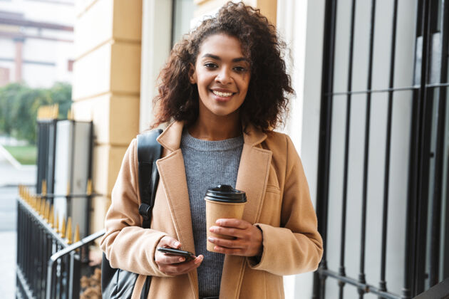 浏览快乐的非洲年轻女子穿着外套走在户外 拿着外卖咖啡杯 用手机非洲休闲街道