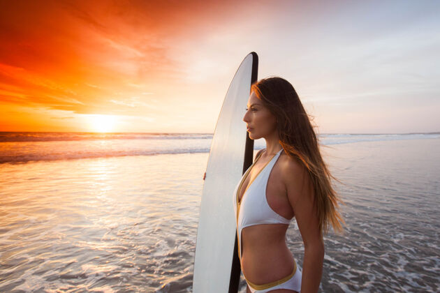 冲浪日落时分 一位穿着比基尼 穿着冲浪板 身材苗条的运动型美女站在海边黎明巴厘岛海洋