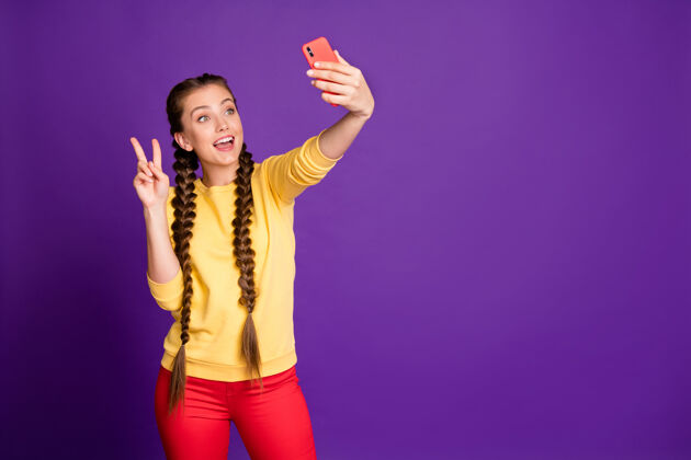 秀漂亮的少女长辫子拿着电话自拍显示v符号穿休闲黄色套头衫红色裤子隔离紫色墙壁自拍手机女性