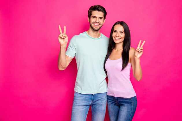 丈夫有趣的夫妇的照片 男男女女举手显示的v型标志说你好和再见穿休闲服隔离亮粉色背景爱情朋友情人