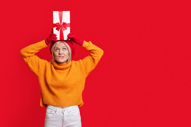 温暖一位金发女郎戴着帽子 穿着暖和的毛衣 头上顶着一个礼品盒 在一个有自由空间的红色工作室墙上摆姿势生日圣诞节梦想