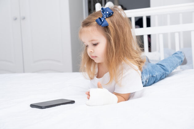 医学手拿石膏的小女孩躺在床上用智能手机 看动画片或教育视频治疗矫形移动