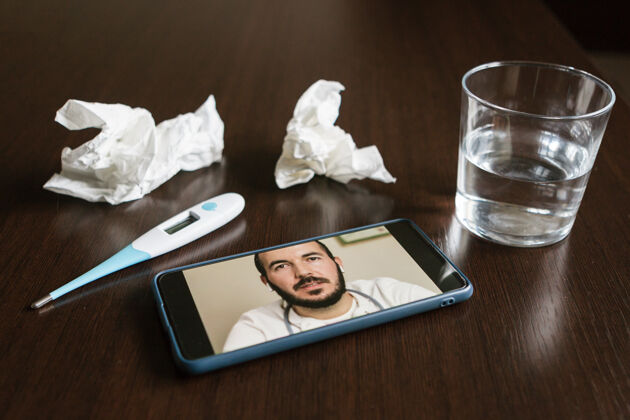 助理手机屏幕上有一位男性医生在线 一些纸巾 一杯水和温度表放在桌子上预约诊断建议