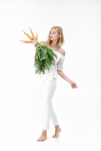 快乐一个穿着白衬衫的金发美女 手里拿着一根新鲜的胡萝卜 上面有绿叶背景.健康还有节食生活方式新鲜微笑