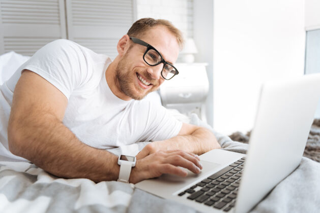 自由职业者愉快的工作时间快乐的微笑胡须男子躺在床上 使用笔记本电脑 同时表达兴趣和工作技术科技访问