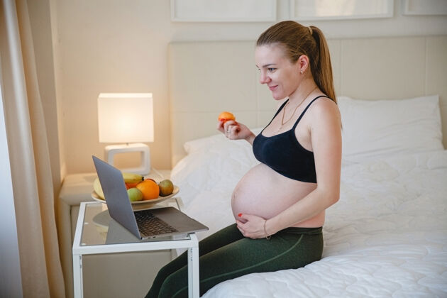 新鲜一个年轻的金发孕妇坐在床上 一边吃水果一边看着她笔记本电脑高高质量的照片期间房间早晨