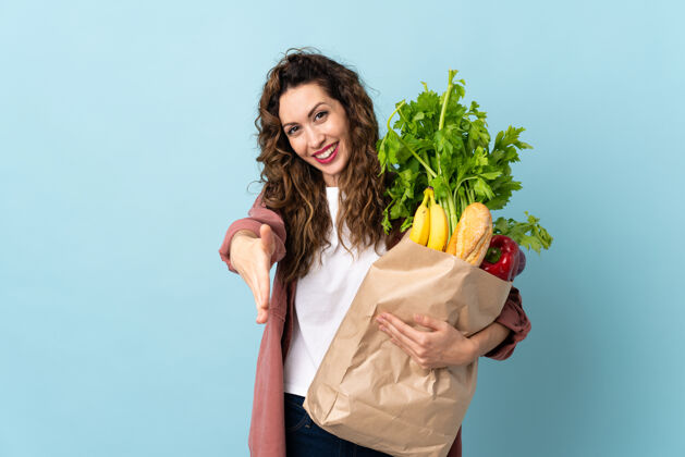 顾客一个年轻的女人拿着一个杂货店的购物袋 孤立地站在蓝色的桌子上 为成交而握手人包蔬菜