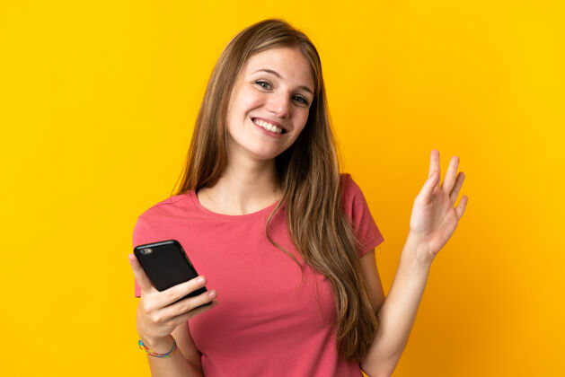 挥手年轻女子用手机隔离在黄色背景上 用手打着招呼 表情愉快你好交谈年轻