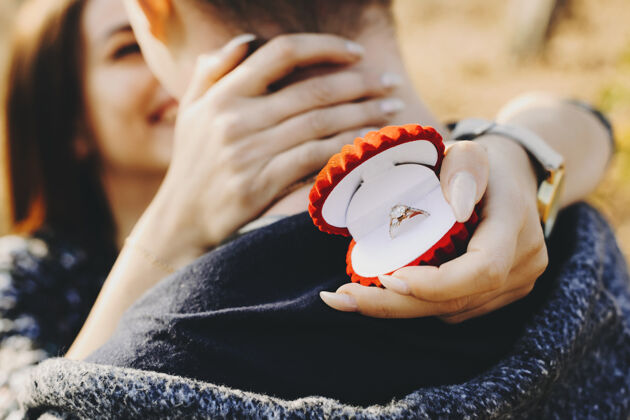 配件在阳光明媚的日子里 年轻的女性微笑着拥抱着无名的男性 手拿着一个戴着优雅订婚戒指的盒子 站在大自然中休闲戒指惊喜