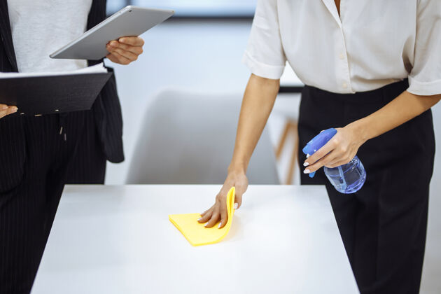 消毒年轻的女商人打扫工作场所 用黄色擦桌子破烂的同事用消毒剂喷雾对工作表面进行消毒 以阻止covid-19扩散工作开放空间女企业