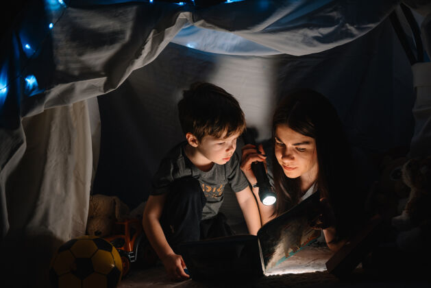 年轻幸福家庭母子俩在帐篷里用手电筒看书主页.家庭概念小故事父母