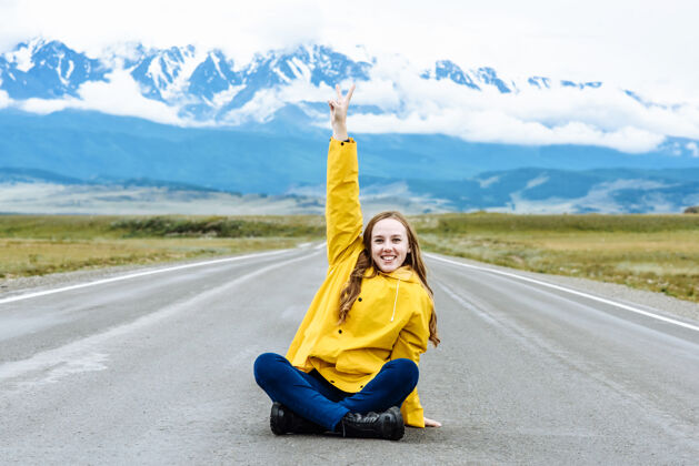 休闲一位年轻开朗的女徒步旅行者面带微笑 坐在雪山背景下的公路上度假 并显示出一种幸福的迹象和平旅行和旅行概念路雪悬崖