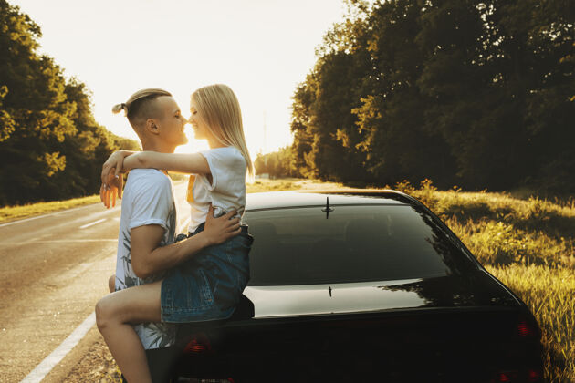 情侣一对令人惊叹的夫妇拥抱的侧视图 而女人坐在路边的车后座上 在夕阳下旅行冒险爱金发