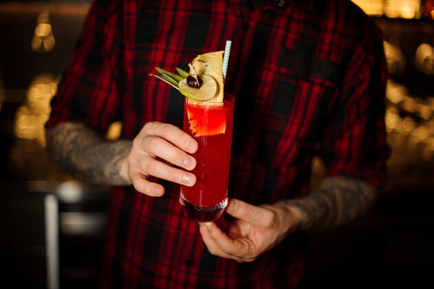 混合有纹身的酒吧男招待拿着一杯红甜多汁饮料 放在一个鸡尾酒杯里 里面有水果强烈餐厅饮料