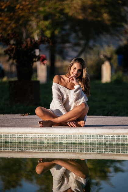 微笑作物赤脚女性盘腿坐在游泳池附近与平静的反射水和微笑在院子里漂亮服装柔软