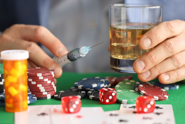 休闲男人的手拿着带针头的注射器和放着酒精的玻璃杯 旁边是赌场筹码赌场俱乐部手