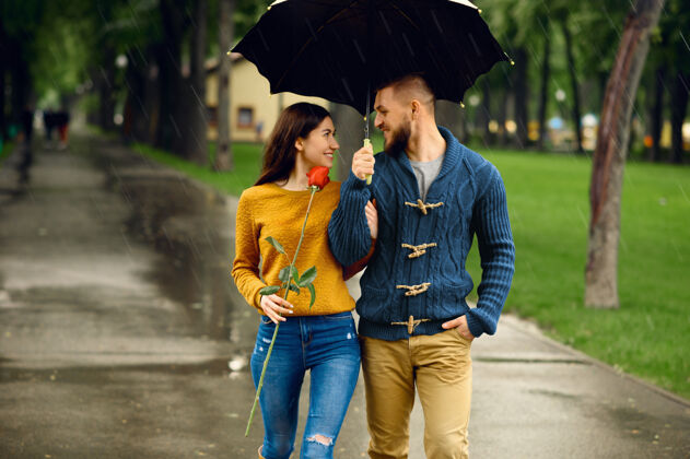 女人雨中带伞的浪漫情侣在夏日公园散步戴曼女人带着玫瑰在雨中走在小路上 雨天在巷子里拥抱一对快乐