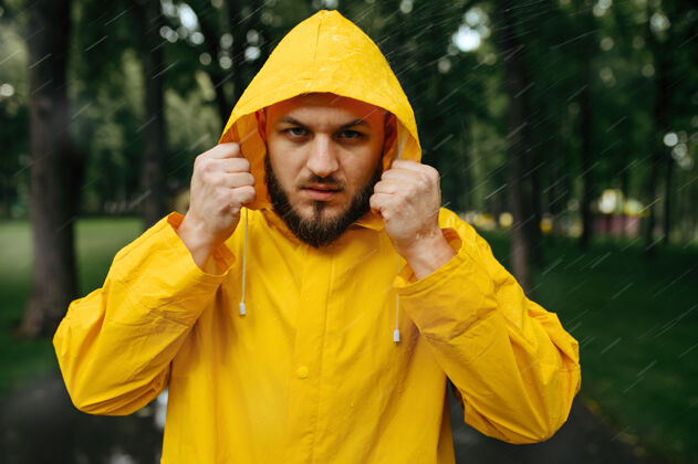 男穿雨衣的人戴上风帽 夏天公园里下雨一天一个人男人穿着雨衣走在小路上 小巷里天气潮湿家伙雨衣保护