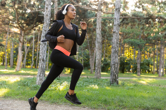运动图为20多岁的运动型女子穿着黑色运动服 戴着耳机 在绿色公园里跑步户外精力充沛减肥