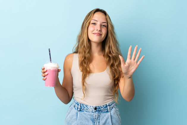 女士拿着草莓奶昔的年轻女子被隔离在蓝色的墙上 用手指数着五个奶昔杯子女孩