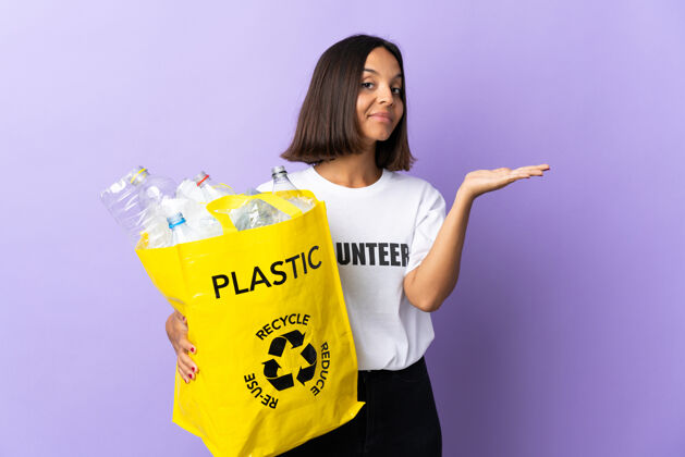 深褐色年轻的拉丁女子拿着一个装满废纸的回收袋 把紫色的手伸到一边 邀请前来回收垃圾手势混合