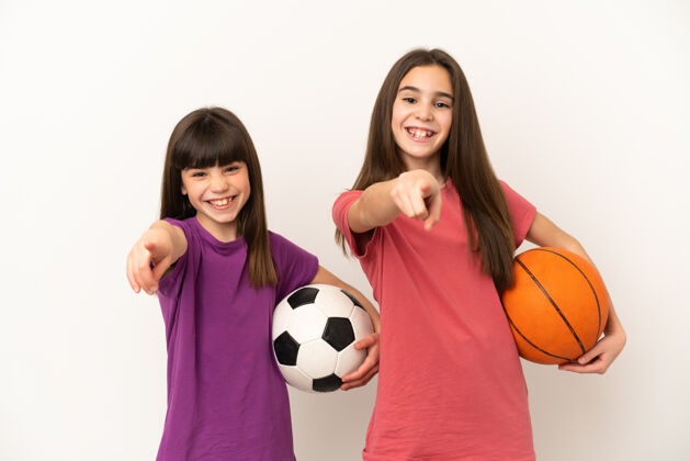 球在白色背景上孤立地踢足球和打篮球的小姐妹们用自信的表情指着你童年信心运动装