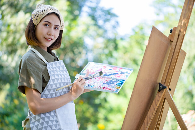 休闲夏日田野里的亚洲美女 用画笔和彩色颜料在花园里的纸艺板上画画女人画笔调色板