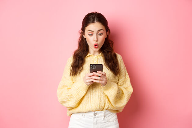 魅力惊讶的年轻女子在智能手机上读短信 说哇哦 好奇地看着手机屏幕 站在粉红色的墙上科技聊天互联网