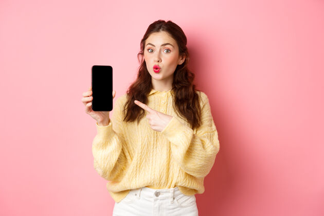 应用程序科技和网上购物兴奋的女孩看起来很好奇 手指指着空手机屏幕 在智能手机上显示应用程序 站在粉红色的墙上应用程序魅力黑发