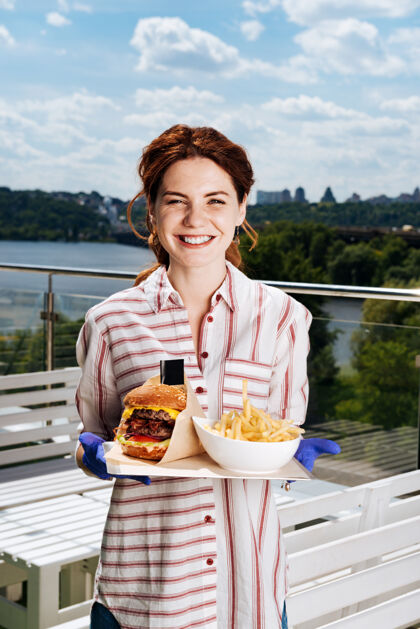 食物大盘喜气洋洋的年轻女子戴着蓝色手套 手里拿着大盘 上面有双层汉堡和朋友土豆快餐服务过程