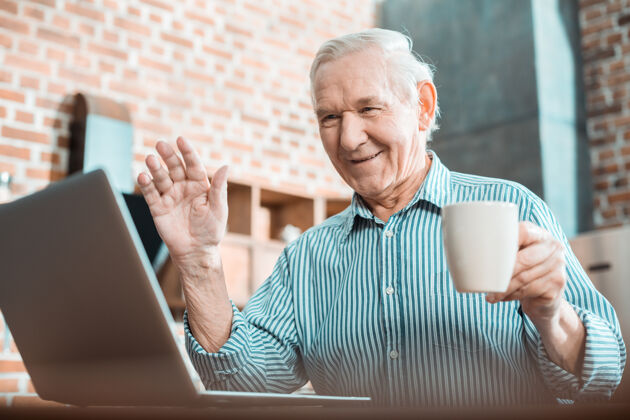 视频一个很好的积极的老人端着一杯茶 一边打视频电话一边打招呼老年人社交互联网