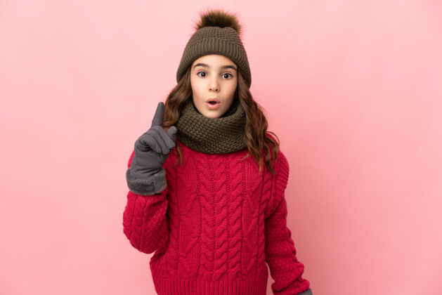 惊喜戴着冬帽的小女孩孤零零地站在粉红色的墙上 一边举起一根手指 一边想实现解决方案温暖沉思女孩