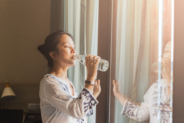 纯净女人站在窗边 在自然光下喝着瓶子里的水科技保健手表