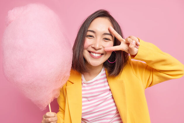 享受漂亮的亚洲年轻女子微笑着积极地做出胜利的姿态眼睛有乐观的心情拿着美味的糖果牙线穿黄色的夹克有甜食的姿势对着粉色的墙壁糖室内棉花