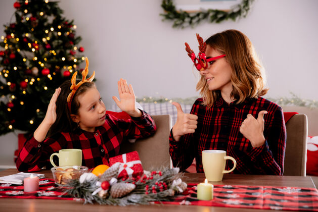 时间戴着驯鹿眼镜的母亲高兴地竖起大拇指看着坐在桌旁的女儿在家里享受圣诞节时光享受桌子坐着